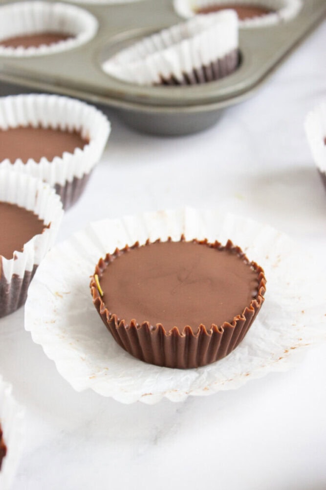 A homemade chocolate peanut butter cup in a brown paper liner sits on a white surface, with a muffin tin and more candy cups in the background.