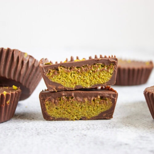 Chocolate cups with a greenish filling are stacked in the center, surrounded by whole chocolate cups on a light surface.