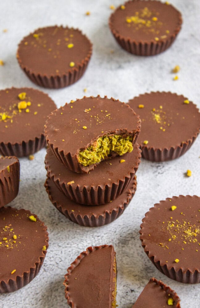 A stack of chocolate cups, one with a bite showing a green filling, surrounded by more chocolate cups on a light surface.