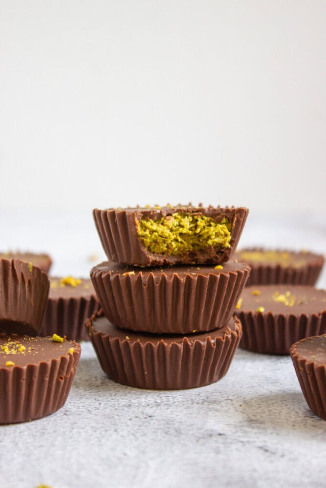 A stack of chocolate cups with a green filling, with the top cup bitten to show the inside, surrounded by more chocolate cups on a light surface.