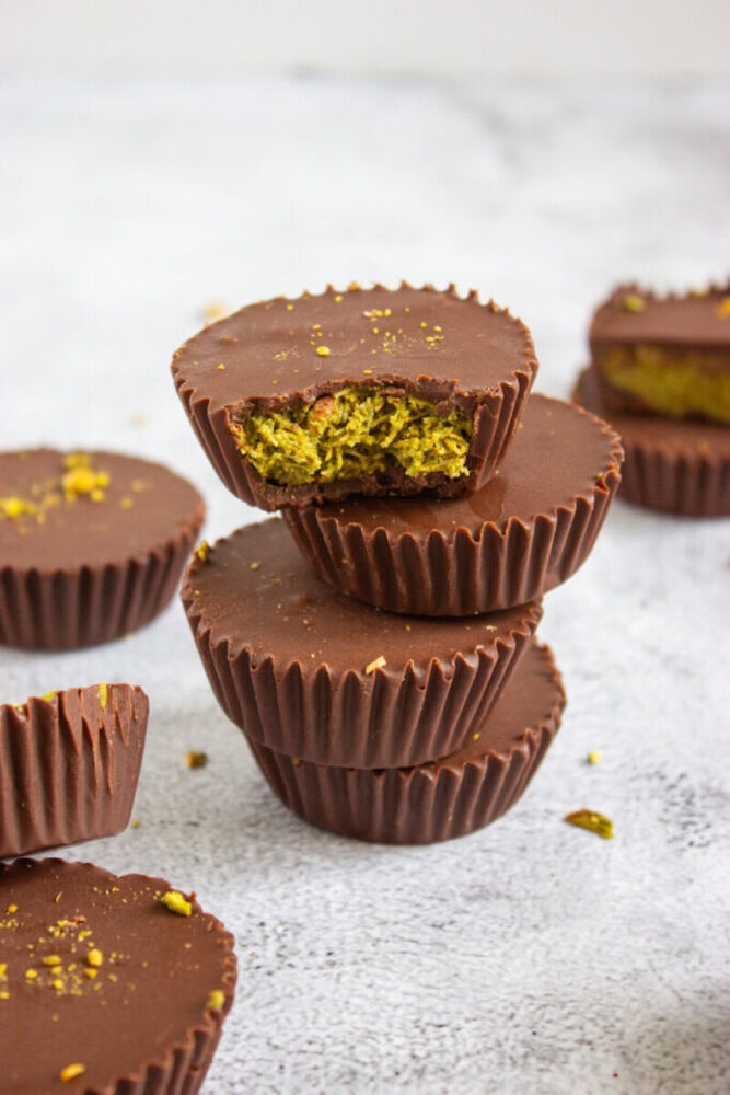 Stack of chocolate cups with a greenish filling, with the top cup bitten to reveal the inside; more cups are scattered in the background on a light surface.