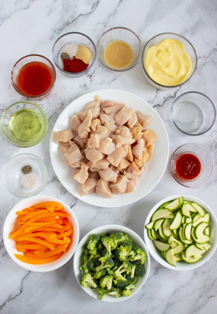 Overhead view of raw diced chicken, sliced zucchini, broccoli, sliced yellow bell pepper, and various sauces and seasonings in bowls on a marble countertop.
