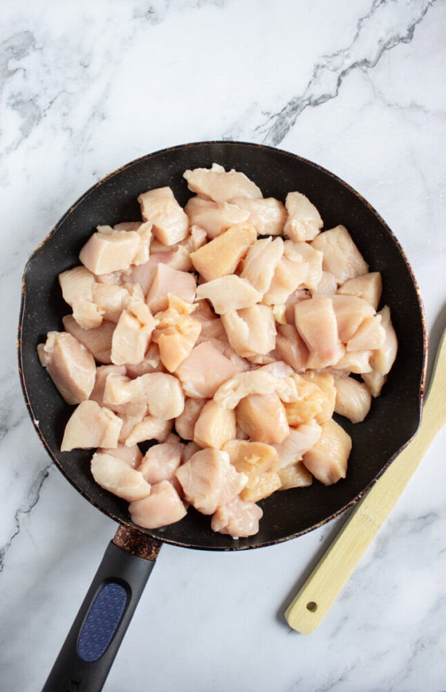 A frying pan filled with raw, diced chicken pieces sits on a marble countertop next to a wooden spatula.