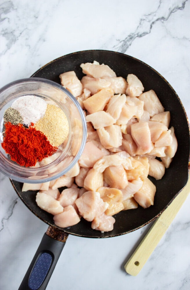 A frying pan with raw diced chicken and a small bowl containing various spices, including paprika, garlic powder, black pepper, and salt, on a marble surface.