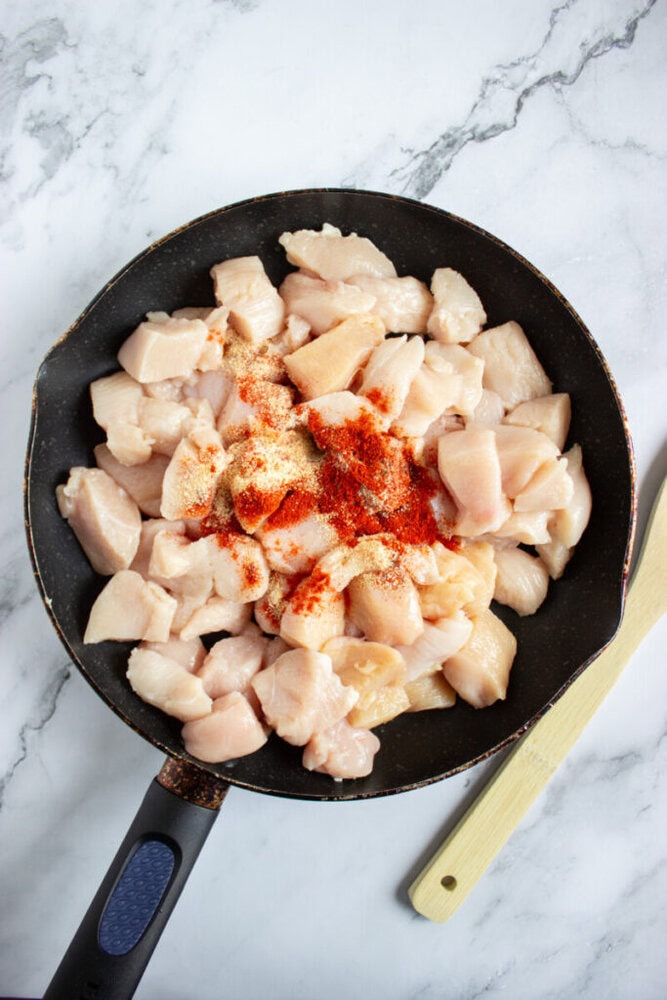 A frying pan with raw, diced chicken breast pieces topped with red and white spices, placed on a marble countertop beside a wooden spatula.