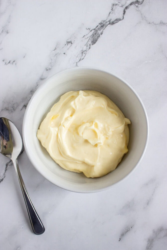 A white bowl filled with creamy mayonnaise sits on a marble surface next to a metal spoon.