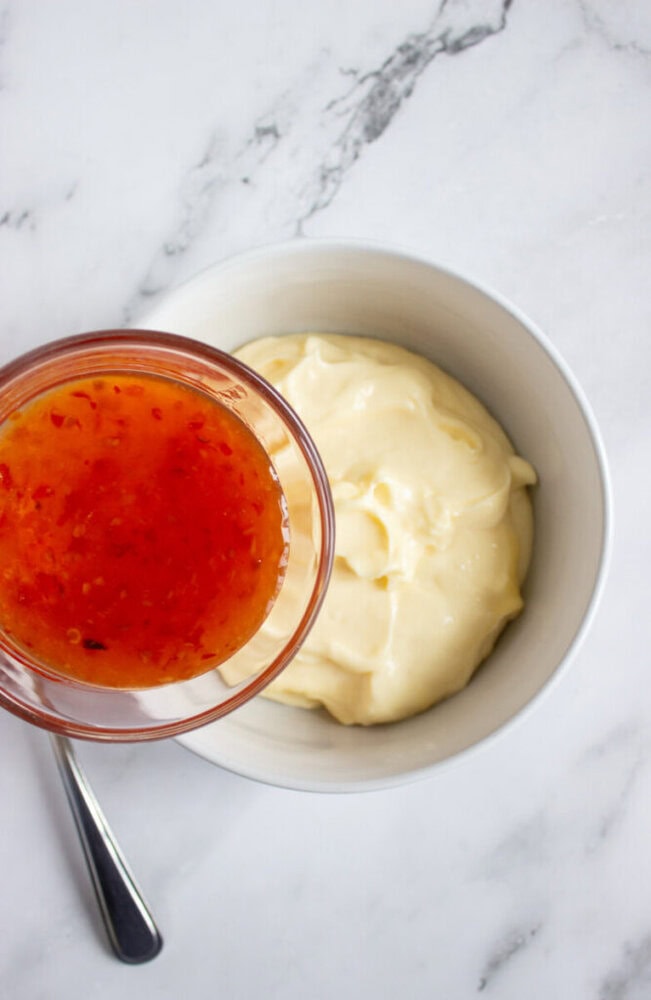 A small glass bowl of sweet chili sauce is held above a white bowl of mayonnaise on a marble surface with a spoon beside the bowls.
