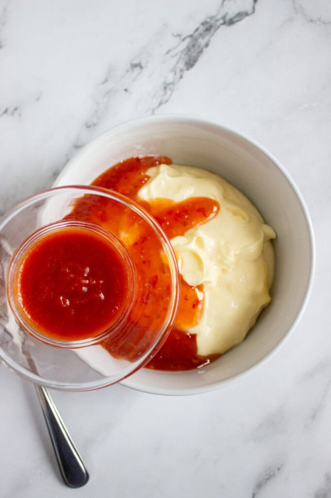 A bowl of mayonnaise with sweet chili sauce being poured on top, set on a white marble surface with a spoon beside the bowl.
