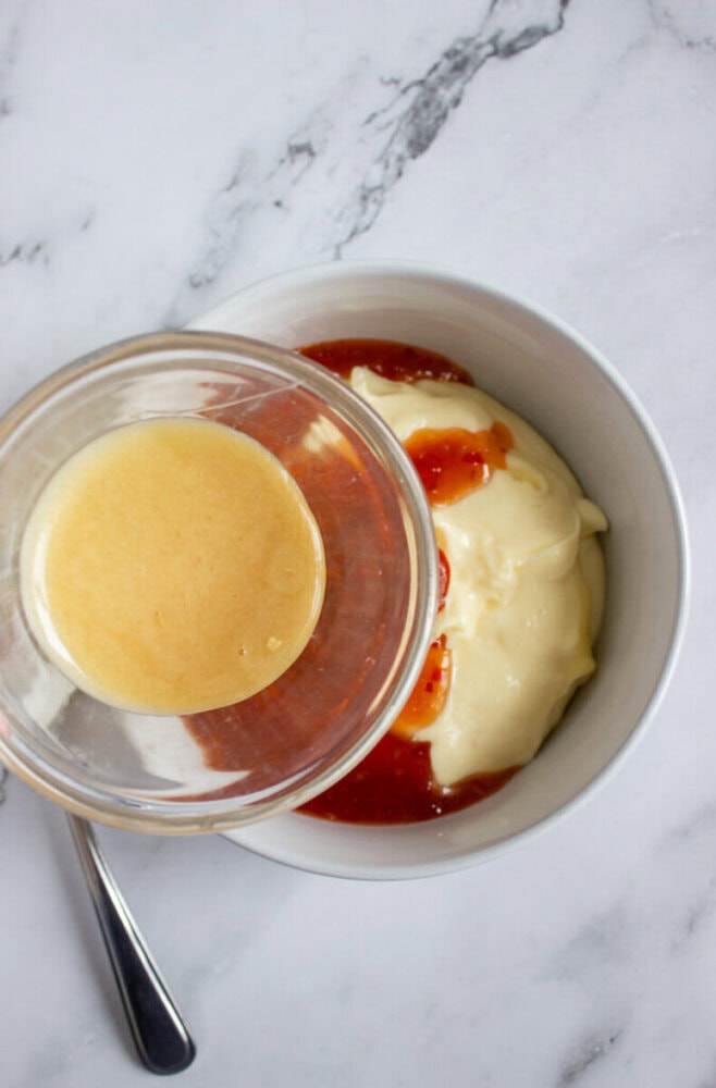 A glass bowl of honey is being poured into a white bowl containing mayonnaise and sweet chili sauce on a marble surface.