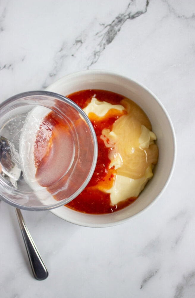 A glass bowl containing mayonnaise, honey, and a red sauce mixture, with a glass of water and a spoon beside it on a white marble surface.