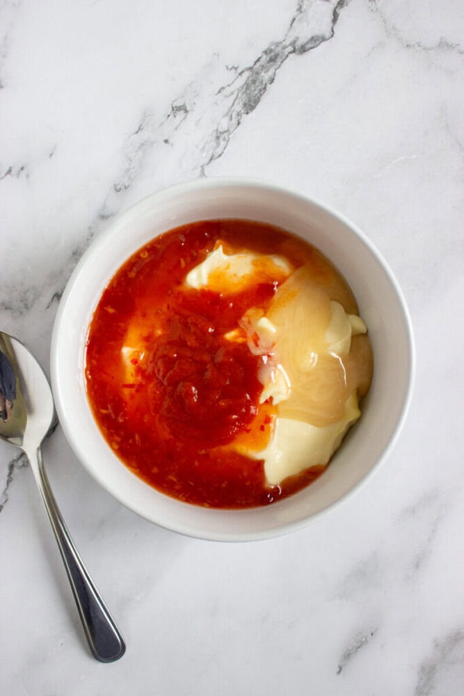 A white bowl containing mayonnaise, sweet chili sauce, and honey, placed on a marble surface next to a metal spoon.