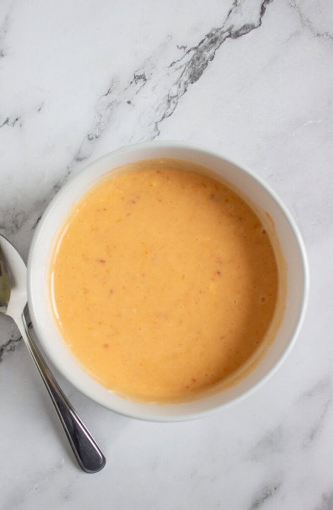A white bowl filled with creamy orange soup or sauce sits on a marble surface next to a spoon.