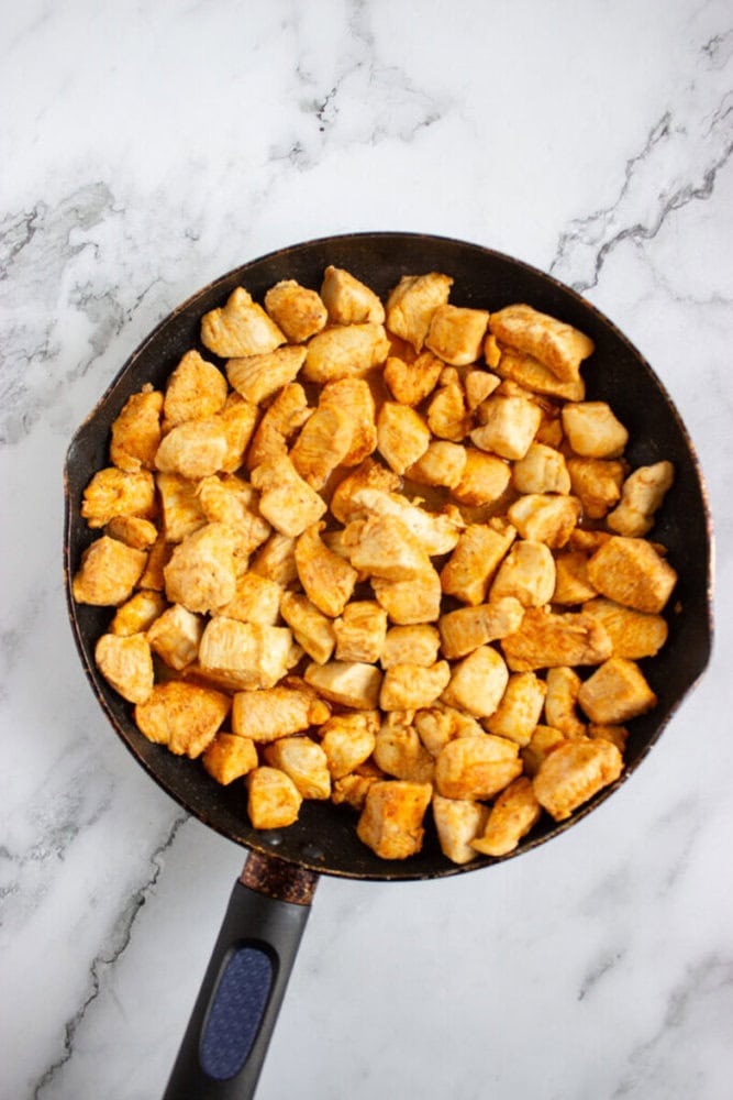 A frying pan filled with cooked, seasoned chicken pieces sits on a white marble surface.