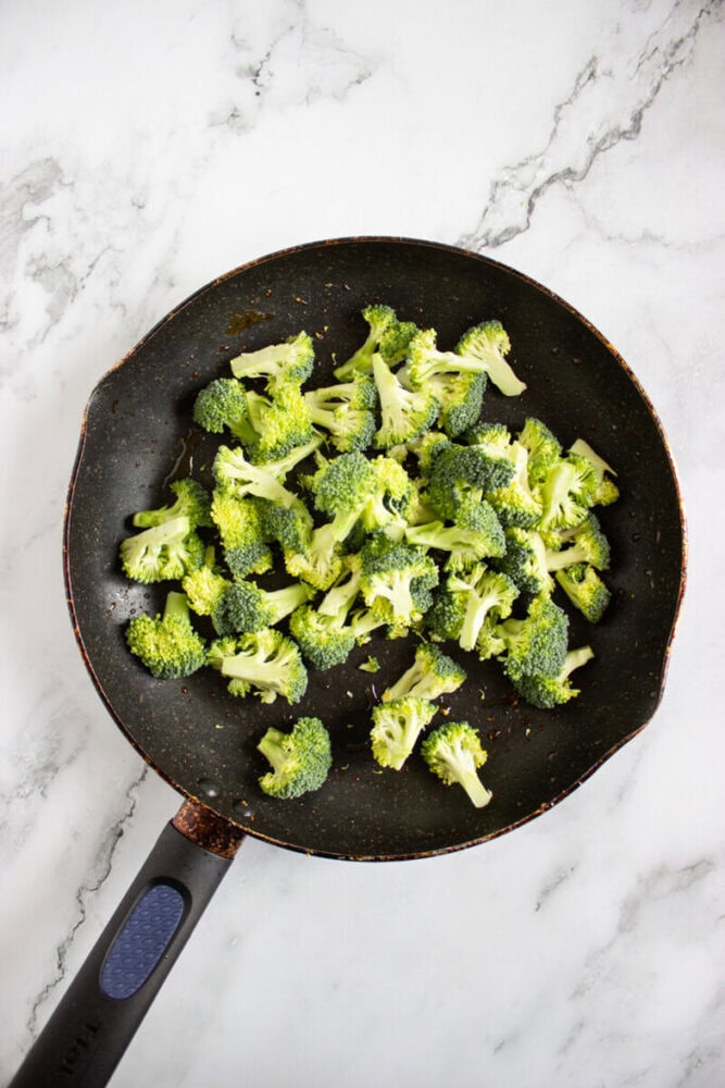 A frying pan with fresh broccoli florets on a marble countertop.