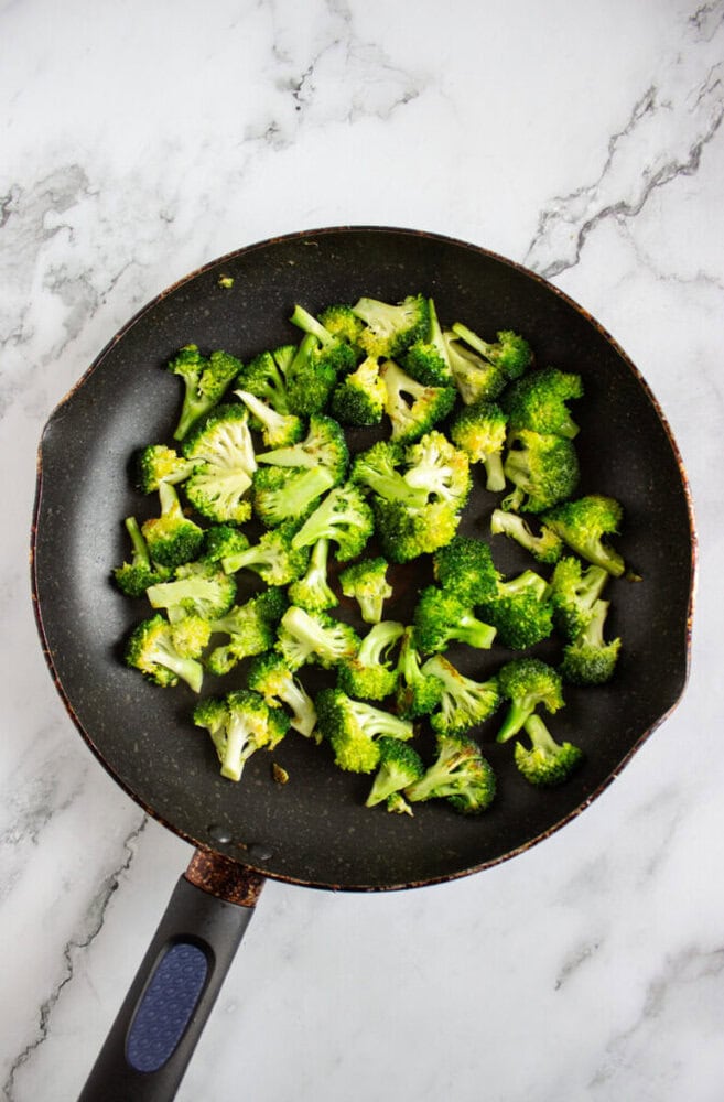 A frying pan with chopped broccoli florets on a marble countertop.