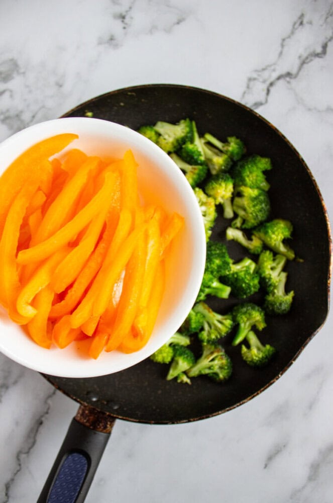A white bowl of sliced yellow bell peppers is being added to a pan of chopped broccoli on a marble countertop.