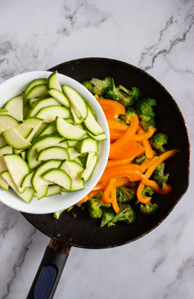 A skillet with broccoli and sliced orange bell peppers, with a bowl of sliced zucchini being poured in, on a marble surface.