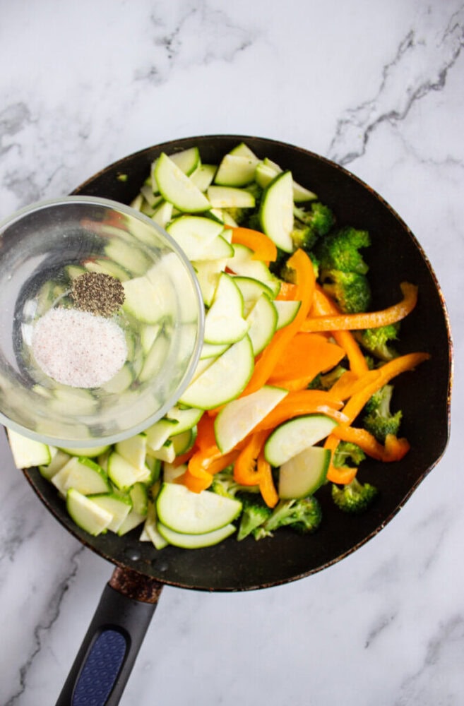 A frying pan with sliced zucchini, yellow bell pepper, broccoli, and a bowl of salt and pepper being added, on a marble countertop.