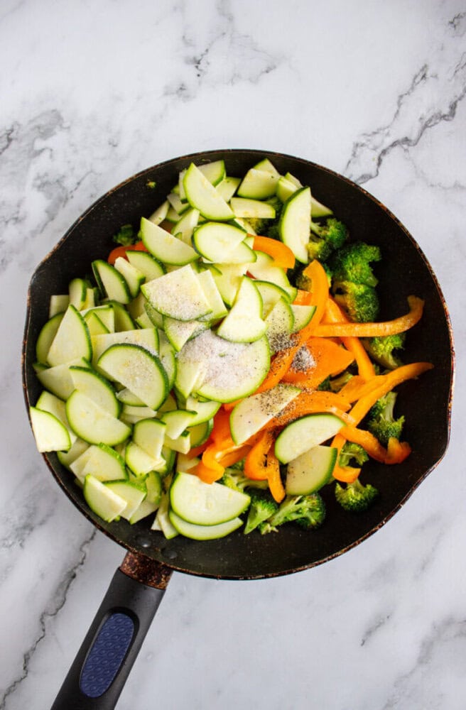 A frying pan on a marble surface contains sliced zucchini, yellow bell pepper strips, and broccoli, seasoned with salt and pepper.