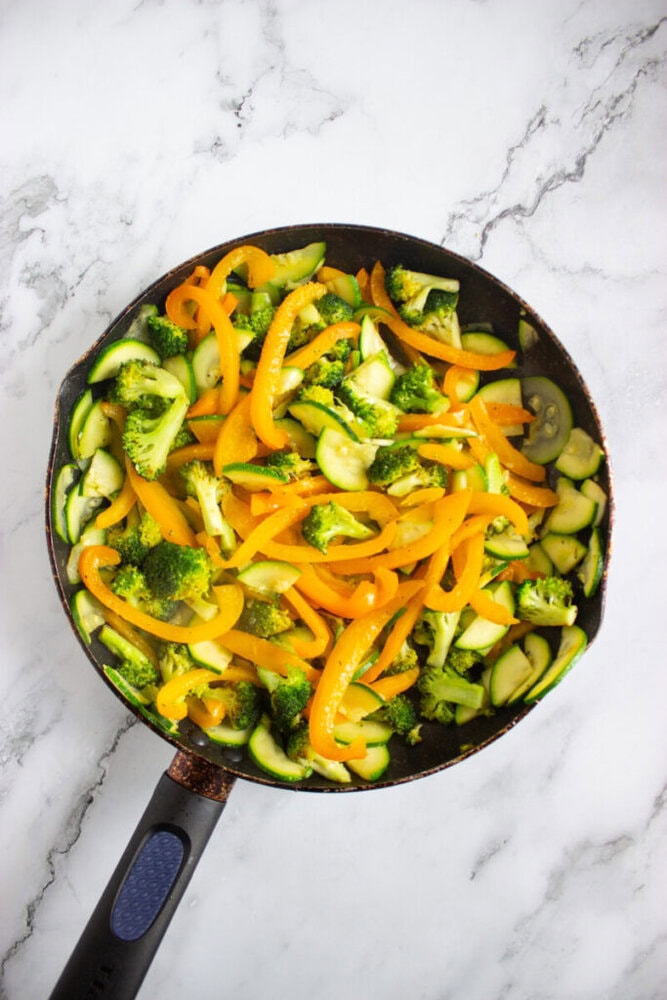 A frying pan filled with sautéed yellow bell peppers, broccoli florets, and sliced zucchini on a marble countertop.