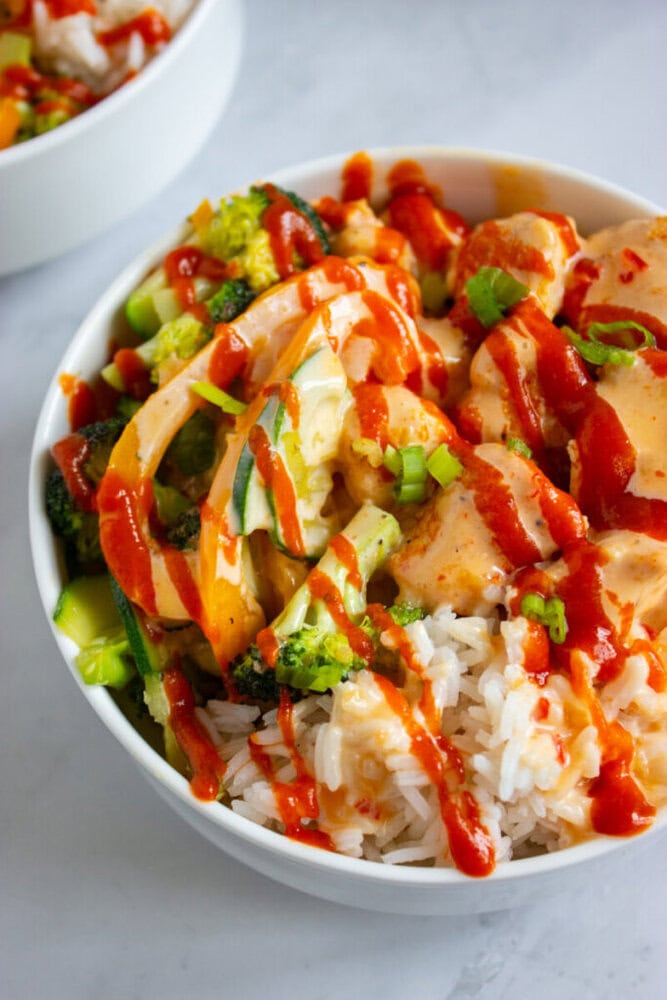 A bowl of white rice topped with chicken, broccoli, sliced green onions, and drizzled with red sauce.
