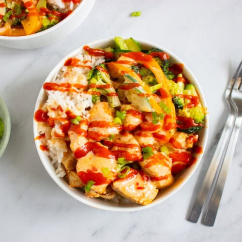 A bowl of rice topped with sliced chicken, colorful vegetables, and drizzled with red sauce, next to a glass bowl of chopped green onions and two forks on a marble surface.