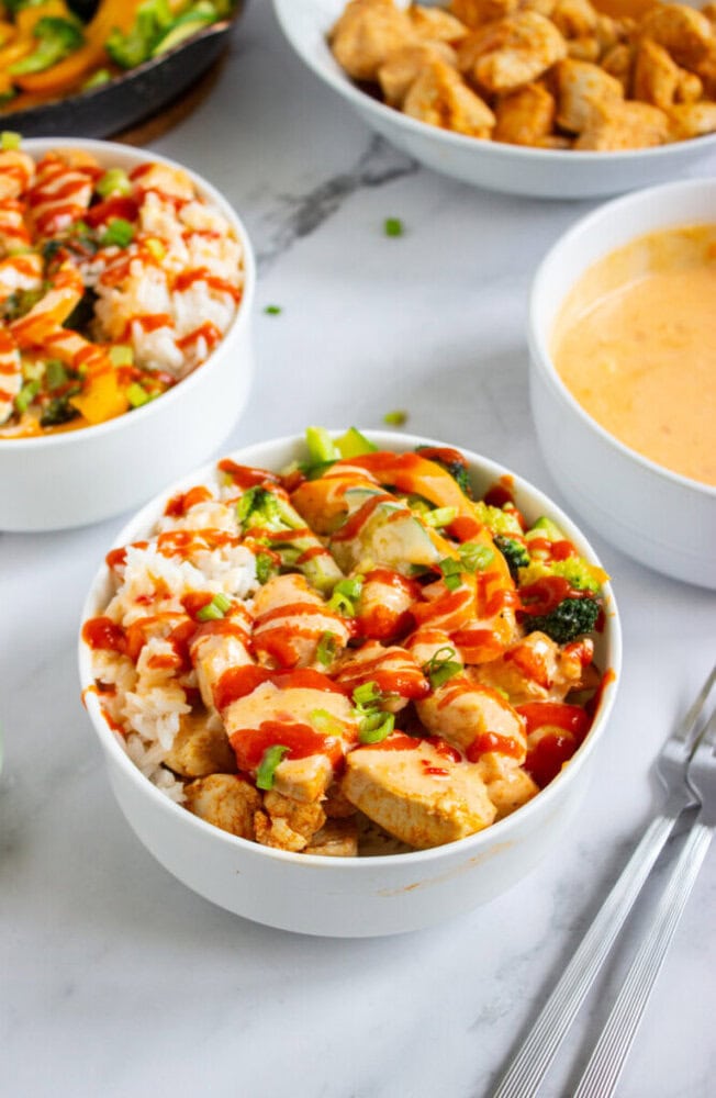 A bowl of rice topped with grilled chicken, broccoli, and vegetables, drizzled with red sauce, with other bowls of food and utensils on a white surface.