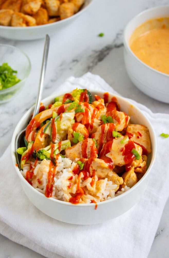 A bowl of white rice topped with chicken, vegetables, green onions, and drizzled with orange sauce and sriracha, with a spoon in the bowl.