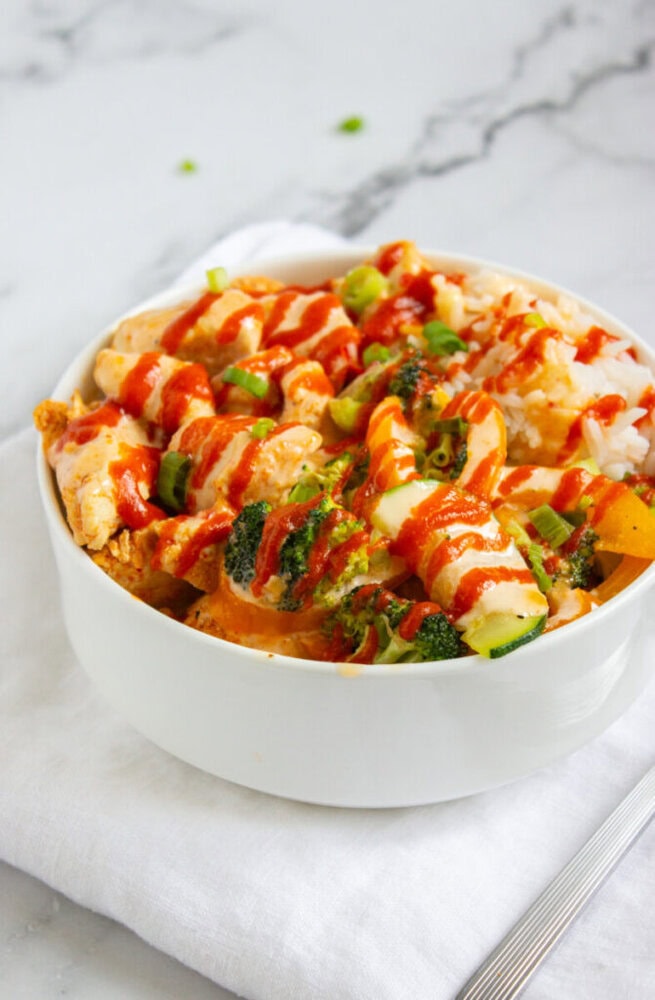 A white bowl filled with rice, chicken, broccoli, and bell peppers, topped with chopped green onions and drizzled with red sauce, placed on a white cloth.