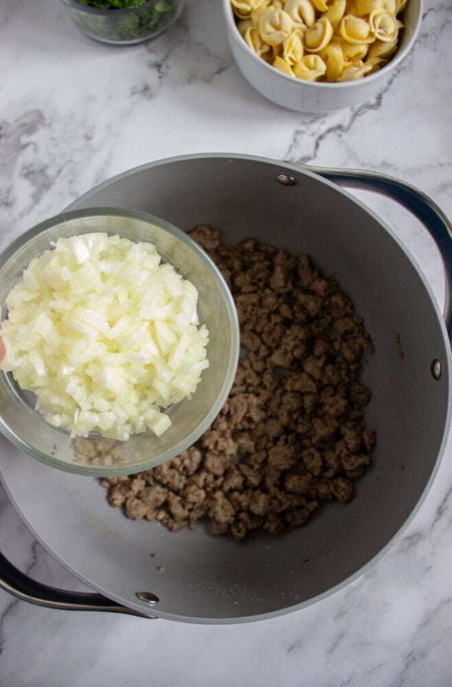 A bowl of chopped onions is being added to a pot with cooked ground meat. Bowls of tortellini and greens are visible in the background on a marble surface.