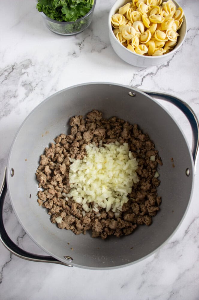 A pot on a marble countertop contains cooked ground meat and chopped onions. Nearby are bowls of fresh tortellini and chopped greens.