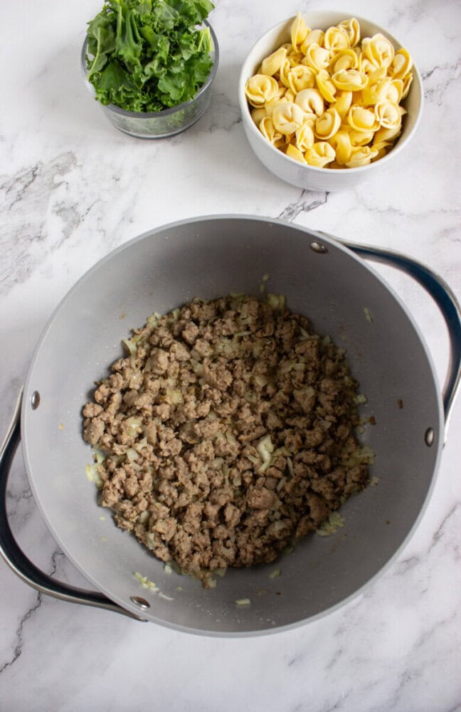 A pot with cooked ground sausage and onions, with bowls of raw tortellini and chopped kale on a marble countertop.