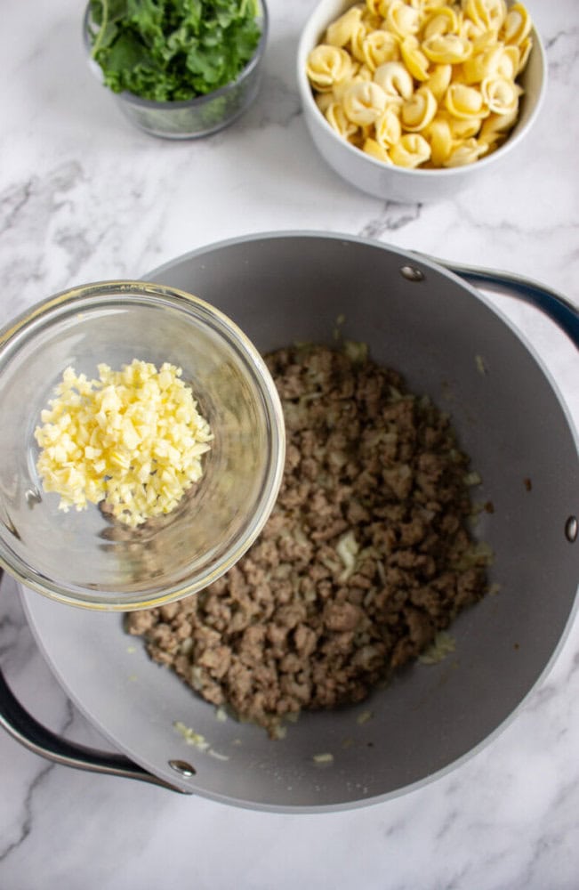 A glass bowl of minced garlic is held above a pot containing cooked ground meat; uncooked tortellini and chopped greens are in bowls nearby.