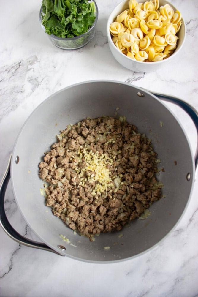 A pot with browned ground meat and chopped garlic, with bowls of fresh spinach and uncooked tortellini on the side, on a marble countertop.