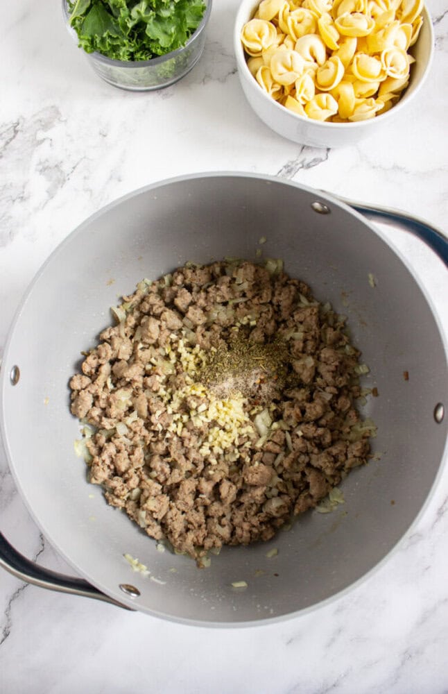 A pot with cooked ground meat, chopped onions, and seasonings. Nearby are bowls of fresh spinach and uncooked tortellini on a marble countertop.