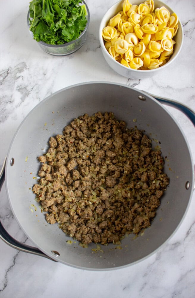 A pot with cooked ground meat and onions sits on a marble countertop next to bowls of fresh spinach and uncooked tortellini pasta.