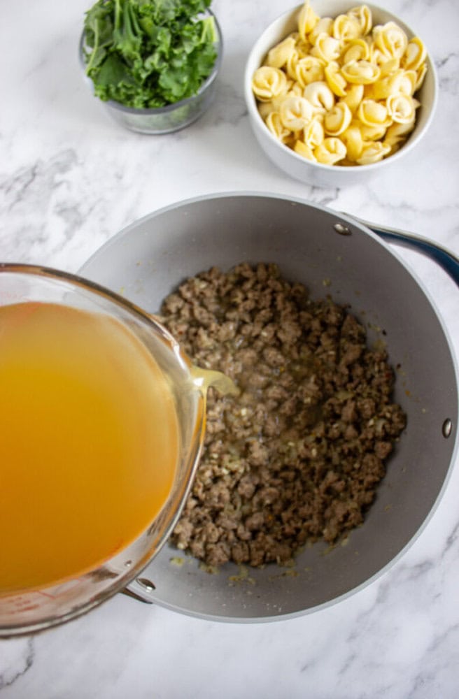 Broth is being poured into a pan with cooked ground meat, with bowls of fresh kale and uncooked tortellini nearby on a marble surface.