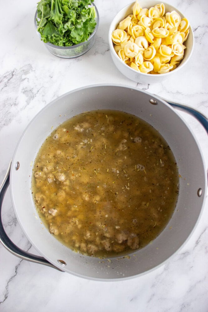 A pot of broth with cooked sausage sits on a marble surface, with bowls of fresh kale and uncooked tortellini placed nearby.