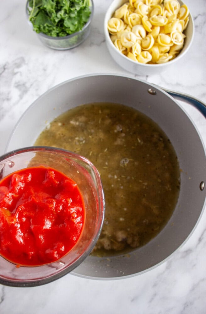 A bowl of diced tomatoes is about to be added to a pot of soup on a marble countertop, with uncooked tortellini and greens in bowls nearby.