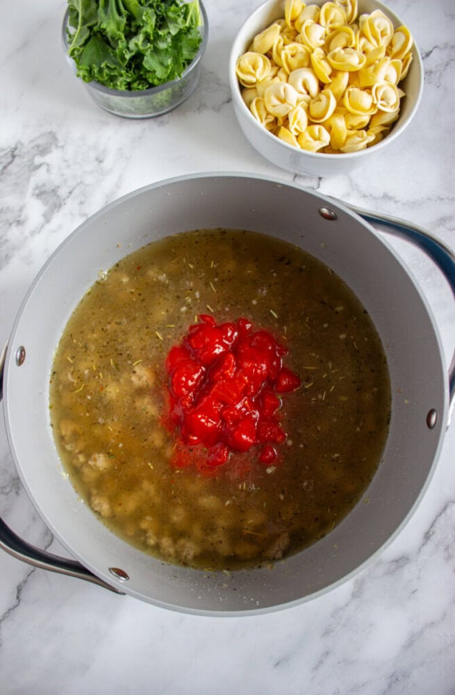 A pot containing broth, seasonings, and diced tomatoes sits on a marble counter, with bowls of fresh kale and uncooked tortellini nearby.
