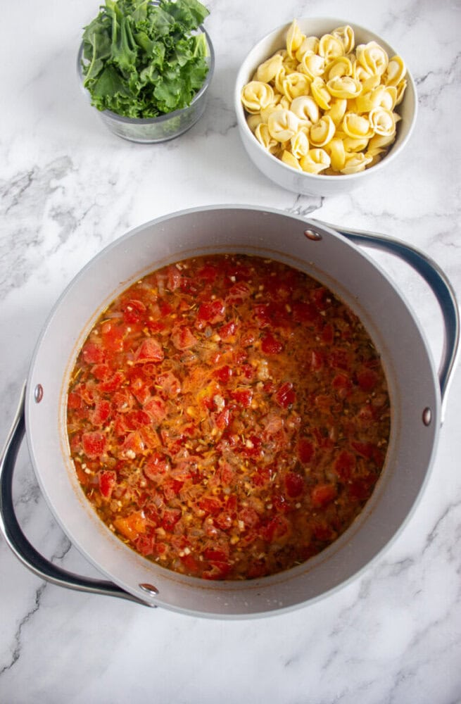 A pot of tomato-based soup simmers on a marble countertop, with bowls of fresh kale and tortellini nearby.