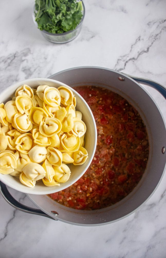 A bowl of uncooked tortellini is being held above a pot of tomato-based soup, with a bowl of leafy greens in the background on a marble countertop.