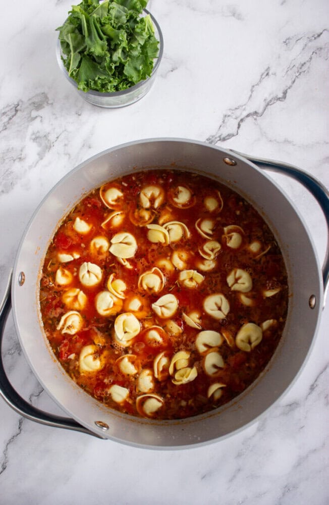 A pot of tomato-based soup with tortellini pieces, next to a bowl of fresh leafy greens, sits on a white marble surface.