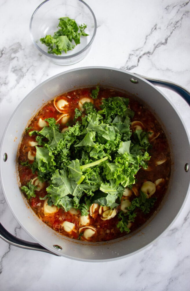 A pot of soup with tortellini, tomato broth, and fresh kale, with a small glass bowl of additional kale nearby on a marble countertop.