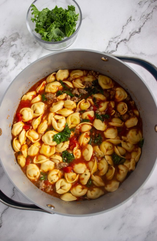 A large pot filled with tortellini soup containing pasta, sausage, tomatoes, and greens sits on a marble surface. A small bowl of fresh parsley is placed nearby.