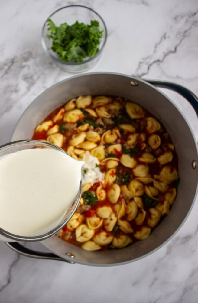 A hand pours cream into a pot of tortellini soup with greens on a marble countertop; a bowl of chopped kale sits nearby.