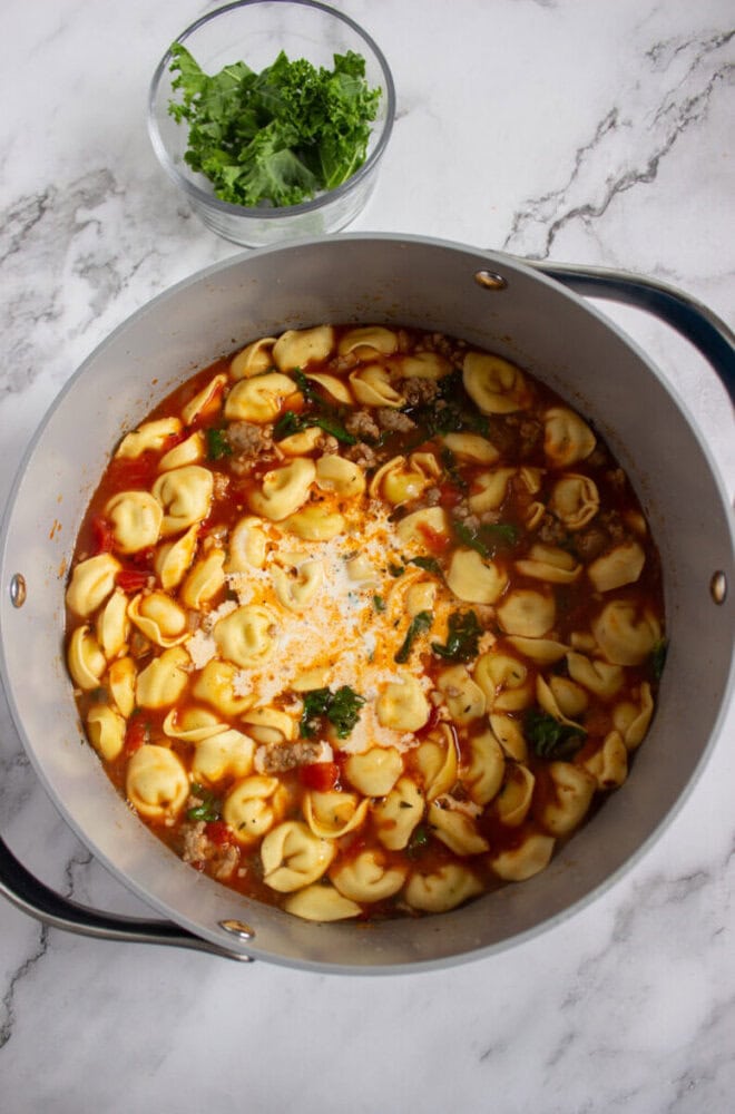 A pot of tortellini soup with tomato broth and greens sits on a marble surface; a small bowl of fresh greens is placed nearby.