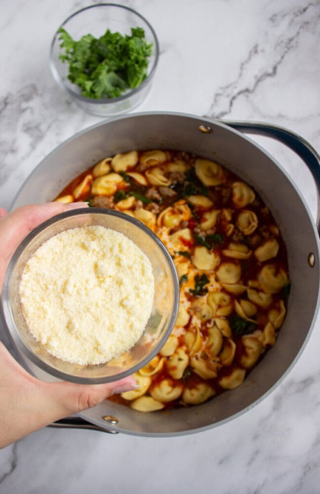 A hand holds a bowl of grated cheese above a pot of tortellini soup on a marble countertop, with a bowl of leafy greens nearby.
