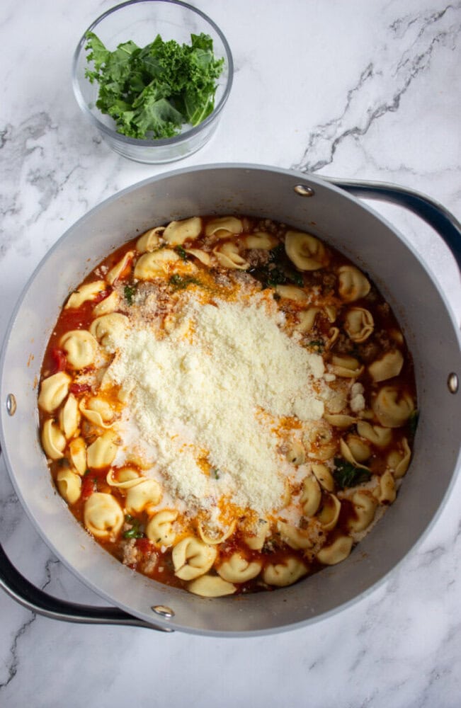 A large pot filled with tortellini soup, tomato broth, greens, and a topping of grated cheese sits on a marble surface; a bowl of fresh kale is nearby.
