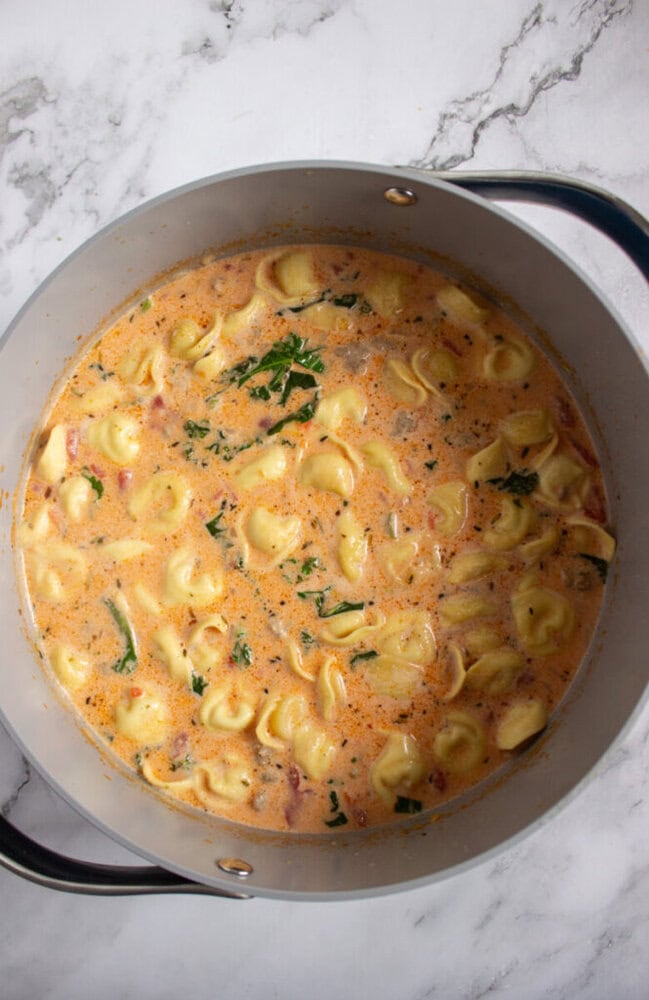 A pot of creamy tortellini soup with herbs and spinach sits on a marble countertop.